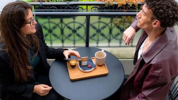 Dos personas sentadas en una mesa redonda con una bandeja que contiene una taza y una caja, al aire libre con una barandilla y plantas al fondo.