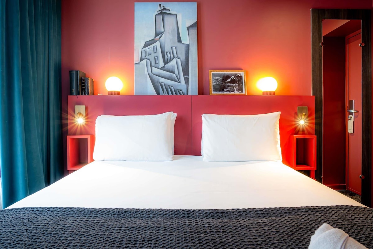 Una habitación de hotel con una cama grande, dos mesitas de noche, un cuadro en la pared y una puerta con picaporte.