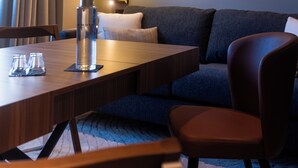 Una habitación de hotel moderna con una mesa de comedor de madera, un sofá y un gran ventanal con cortinas.