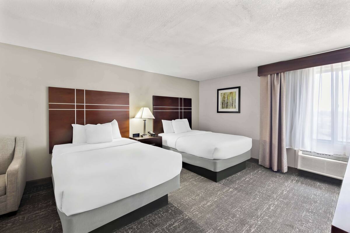 Une chambre d’hôtel avec deux lits, un bureau, une chaise, une lampe, un tableau et une fenêtre avec des rideaux.