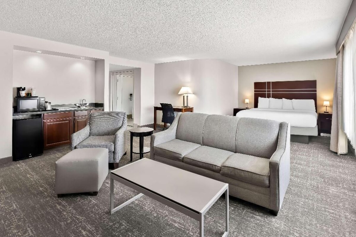 A hotel room with a sofa, a small table, a chair, a bed, a desk with a lamp, and a kitchenette.