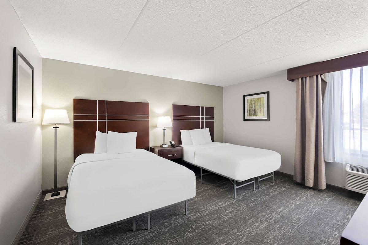 Une chambre d’hôtel avec deux lits, une table de chevet avec une lampe, un tableau encadré au mur et une fenêtre avec des rideaux.