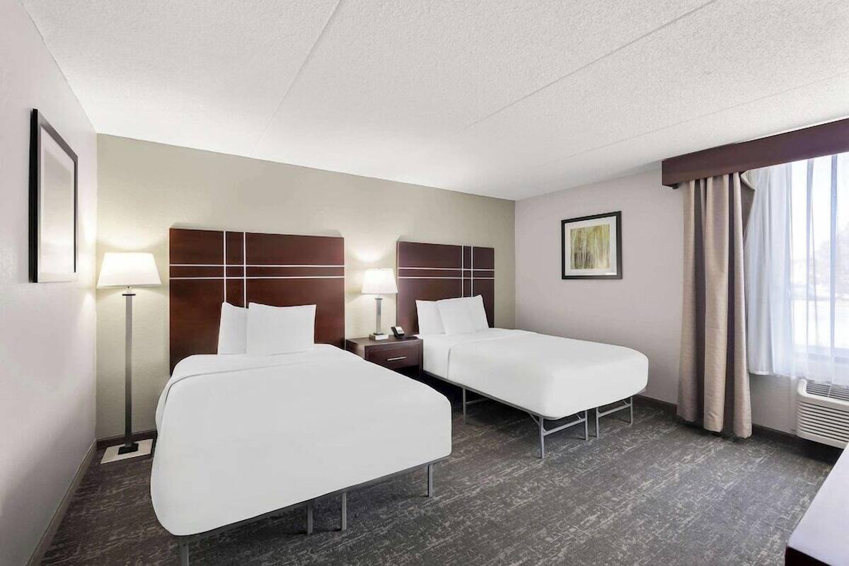 Une chambre d’hôtel avec deux lits, une table de chevet, une lampe, un tableau encadré au mur et une fenêtre avec des rideaux.
