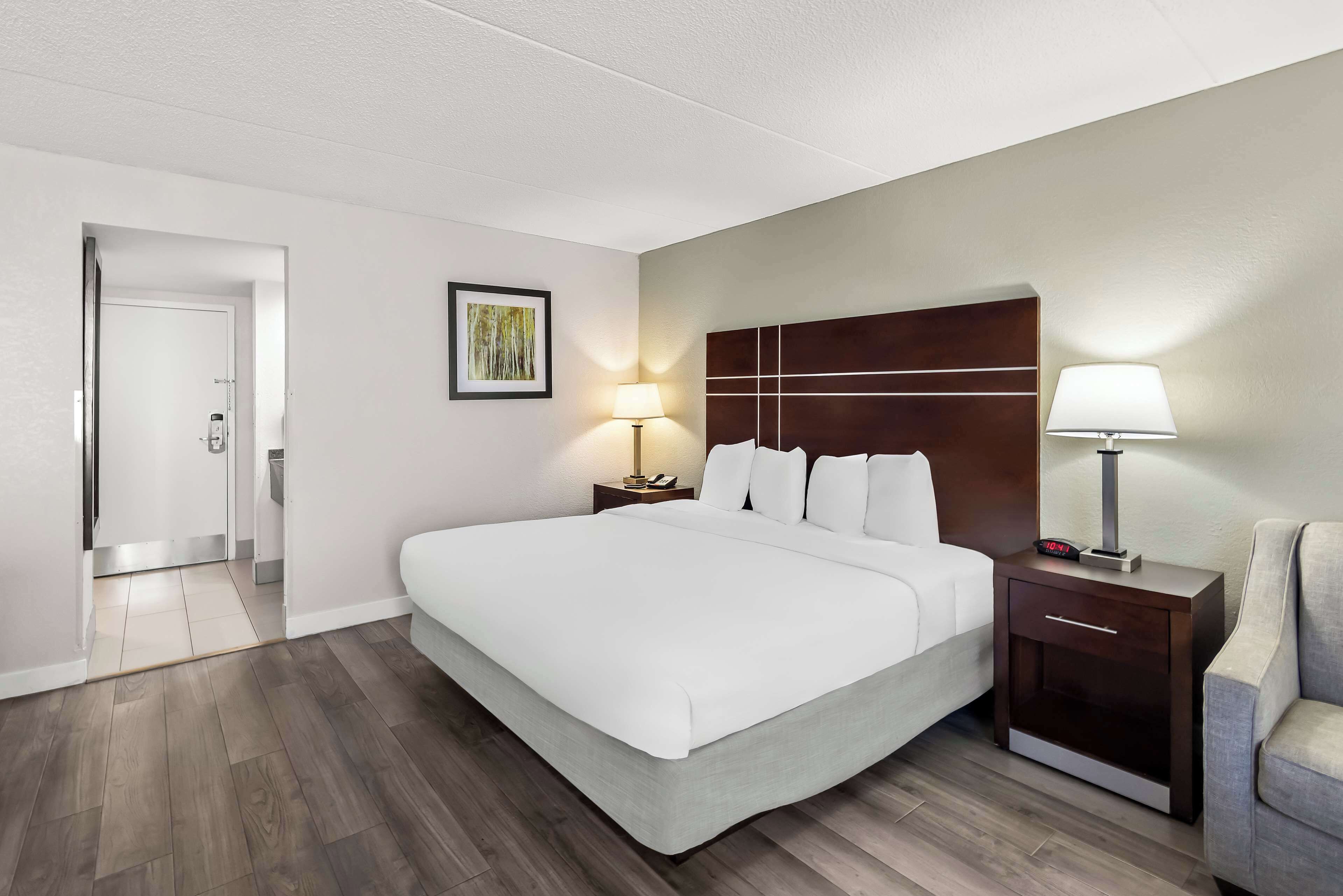 Habitación de hotel con una cama grande, una mesita de noche con lámpara, una silla y una puerta que da a otra habitación.