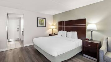 A hotel room with a large bed, a nightstand with a lamp, a chair, and a door leading to another room.