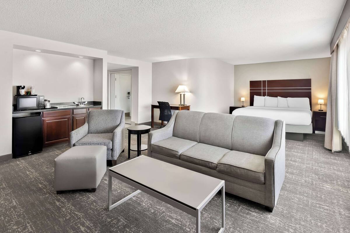 Une chambre d’hôtel avec un canapé, une petite table, une télévision, un coin cuisine et un lit avec une tête de lit.
