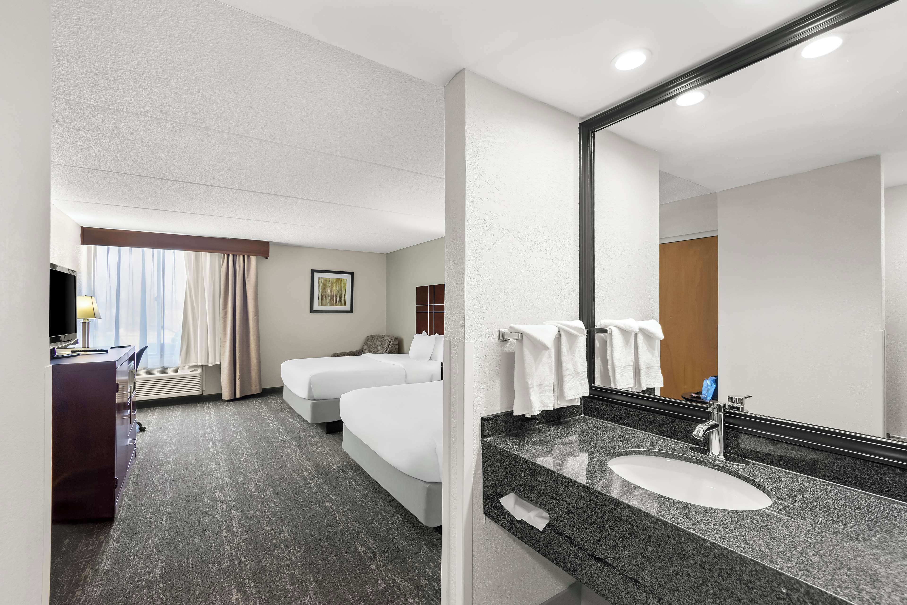 A hotel room with a large bed, a desk with a lamp, a bathroom with a sink and mirror, and a window with curtains.
