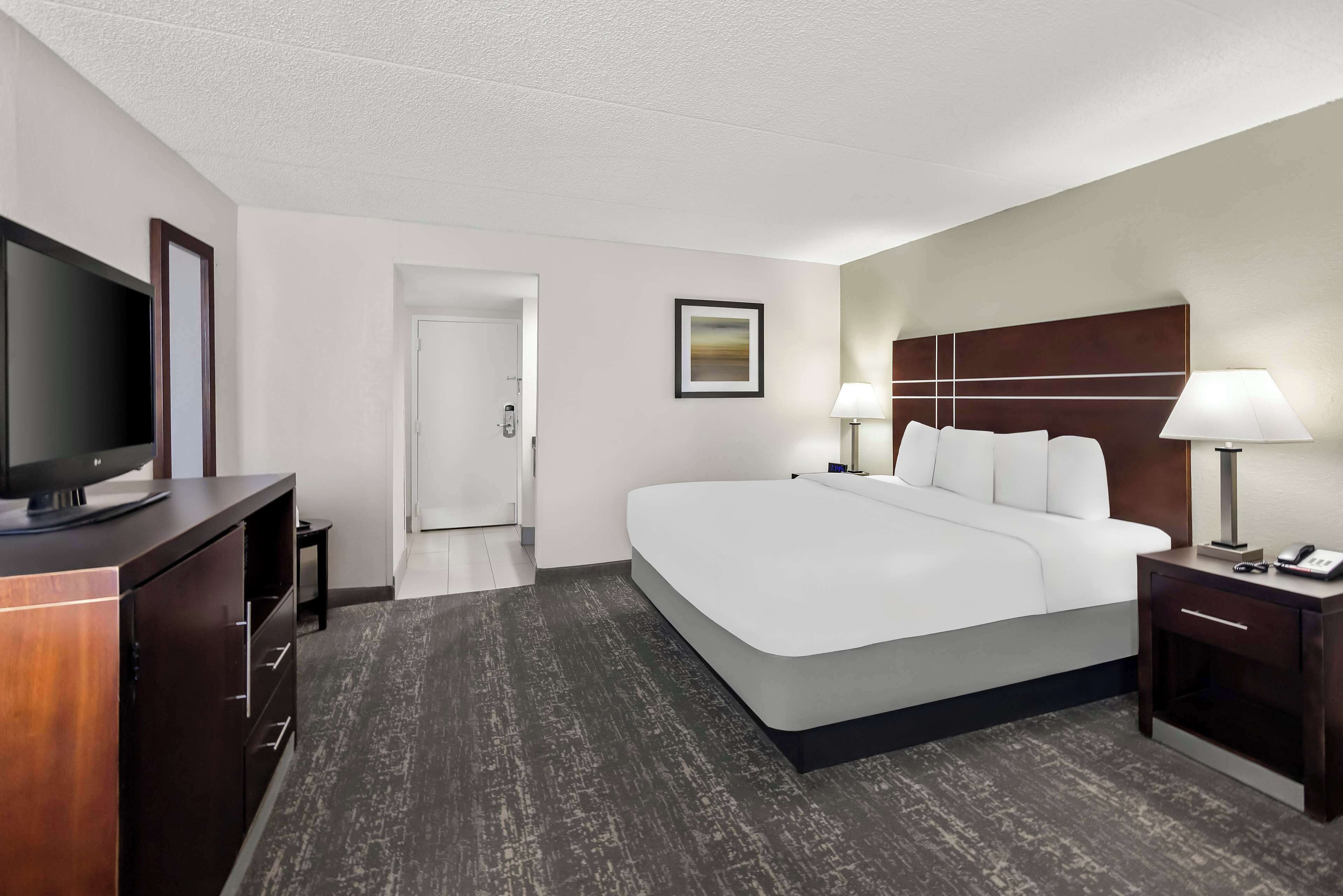 A hotel room with a large bed, a TV on a wooden cabinet, a bedside table with a lamp, and a door leading to a bathroom.