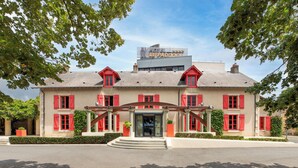 A two-story building with a sign reading "Hotel Le Paddock." It features a central entrance with steps, red shutters, and a balcony on the second floor.