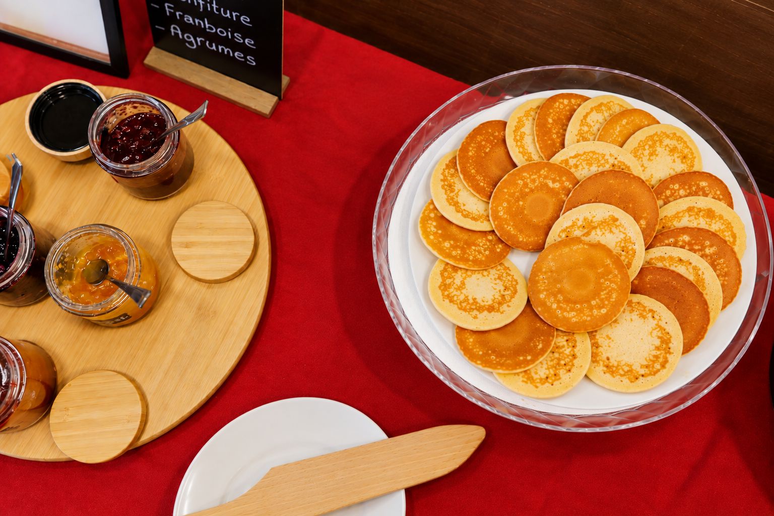 Une table dressée pour le petit-déjeuner, avec des crêpes et des confitures sur une nappe rouge.