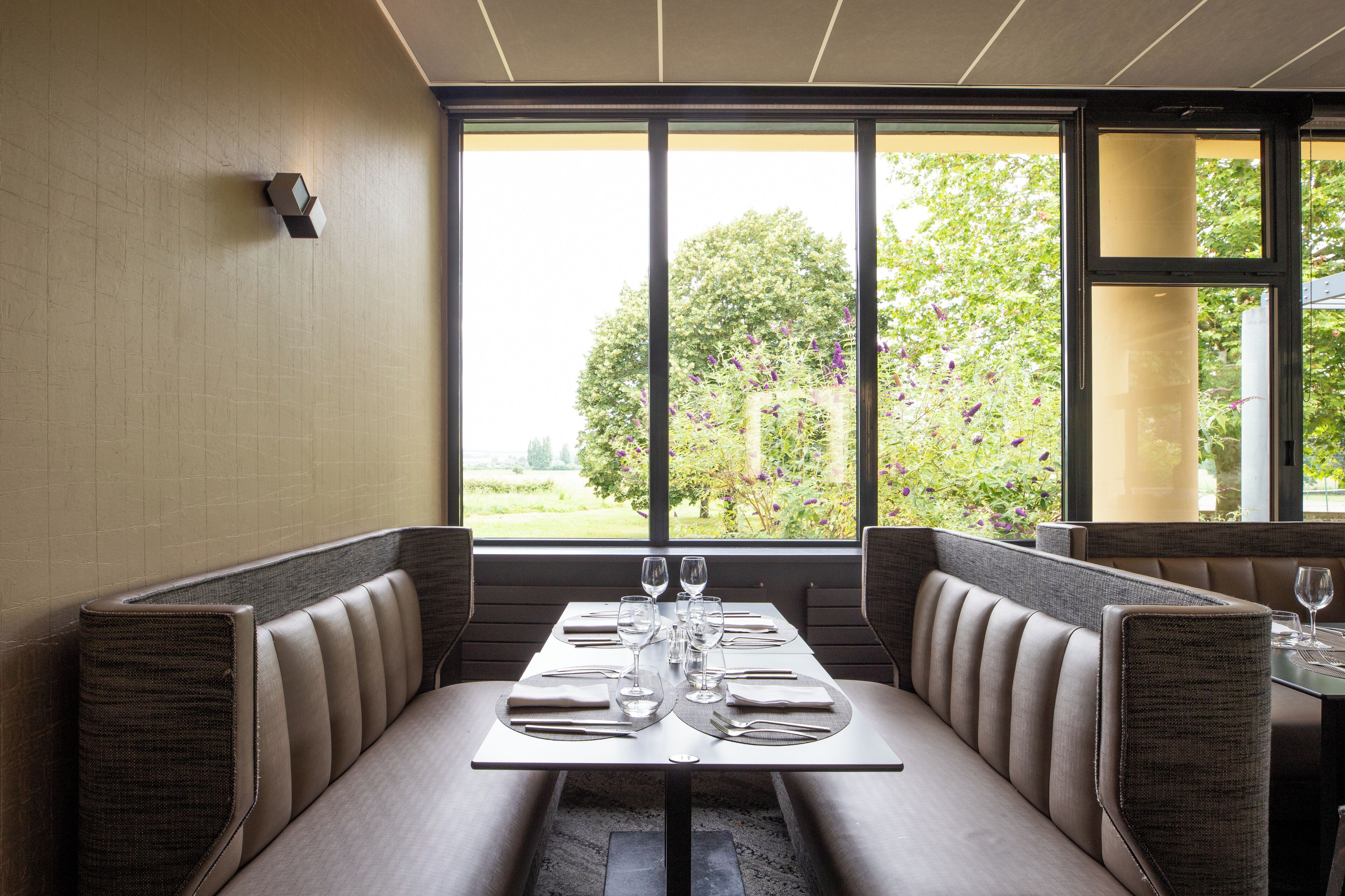 Un restaurant avec des banquettes, une table dressée avec des verres à vin et des assiettes, et une vue sur la verdure à l’extérieur.