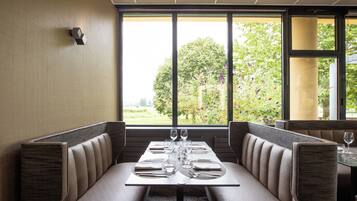Un restaurant avec des banquettes, une table dressée avec des verres à vin et des assiettes, et une vue sur la verdure à l’extérieur.