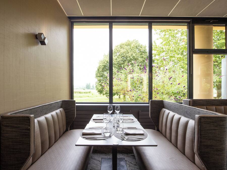Un restaurant avec des banquettes, une table dressée avec des verres à vin et des assiettes, et une vue sur la verdure à l’extérieur.