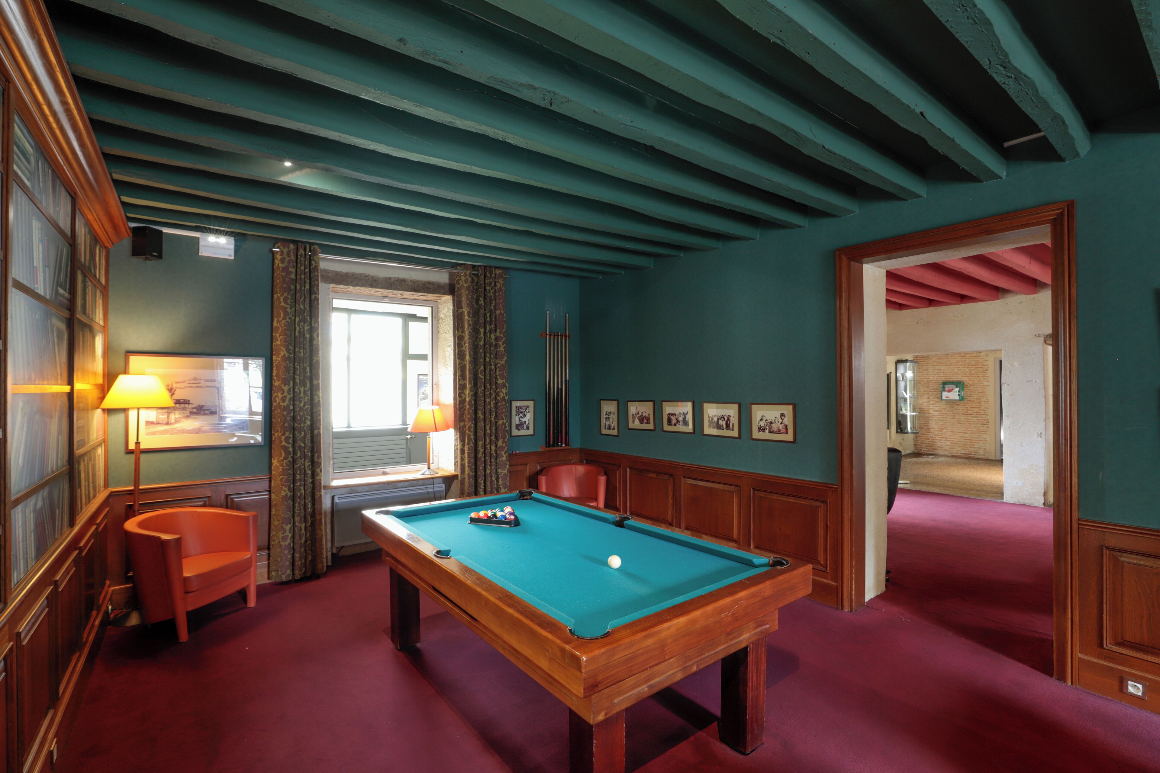 A pool table in a room with teal walls and wooden ceiling beams, a red carpet, and framed pictures on the wall.
