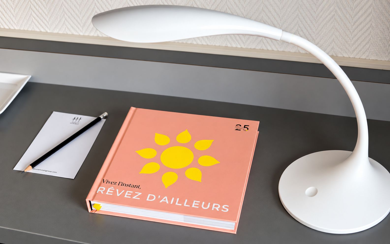 A desk with a book titled 'Vivez l'instant, REVEZ D'AILLEURS,' a pen, and a white desk lamp.