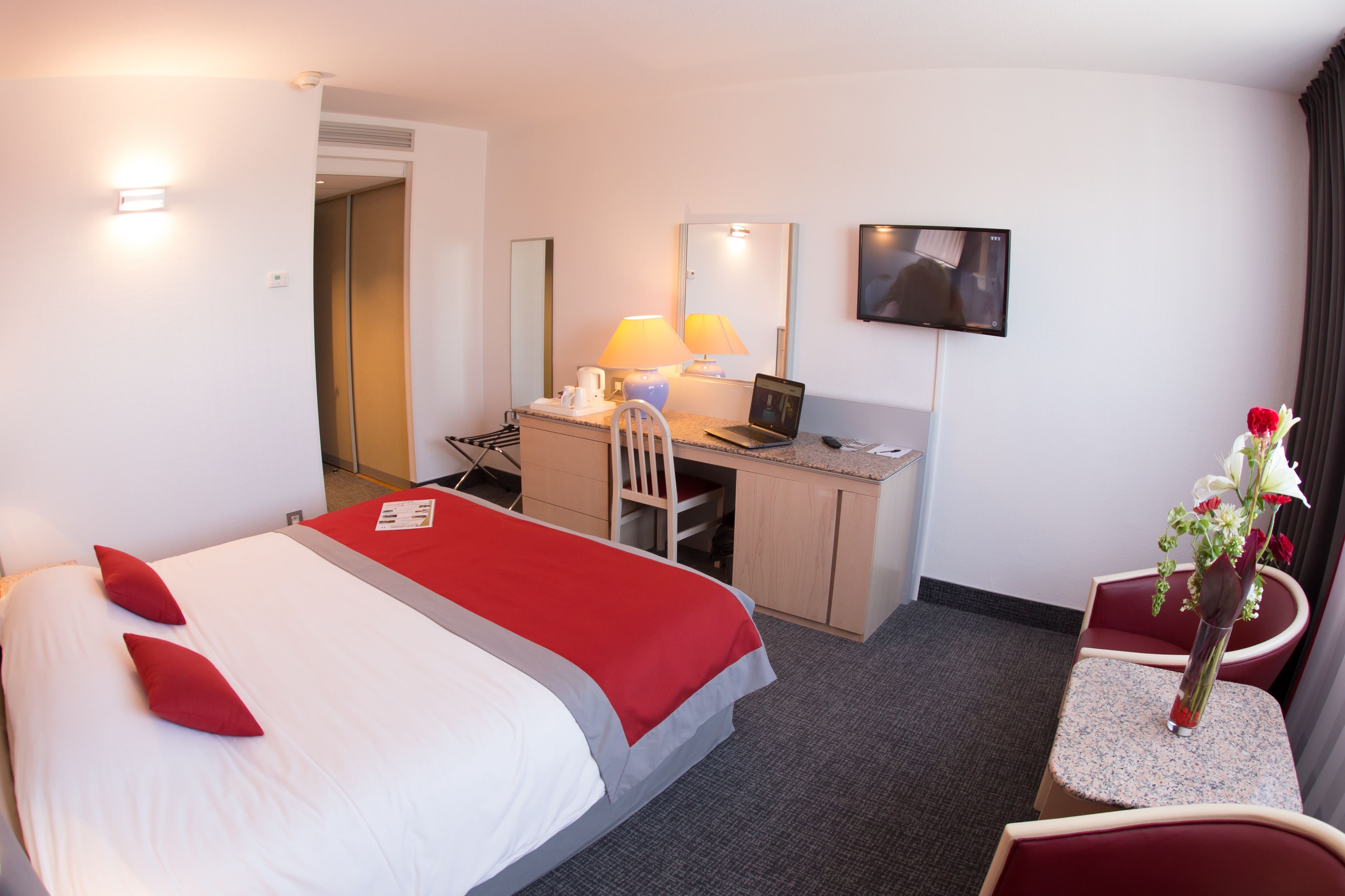 Ein Hotelzimmer mit Bett, Schreibtisch, Stuhl, Laptop, Fernseher und einem kleinen Tisch mit Blumen.