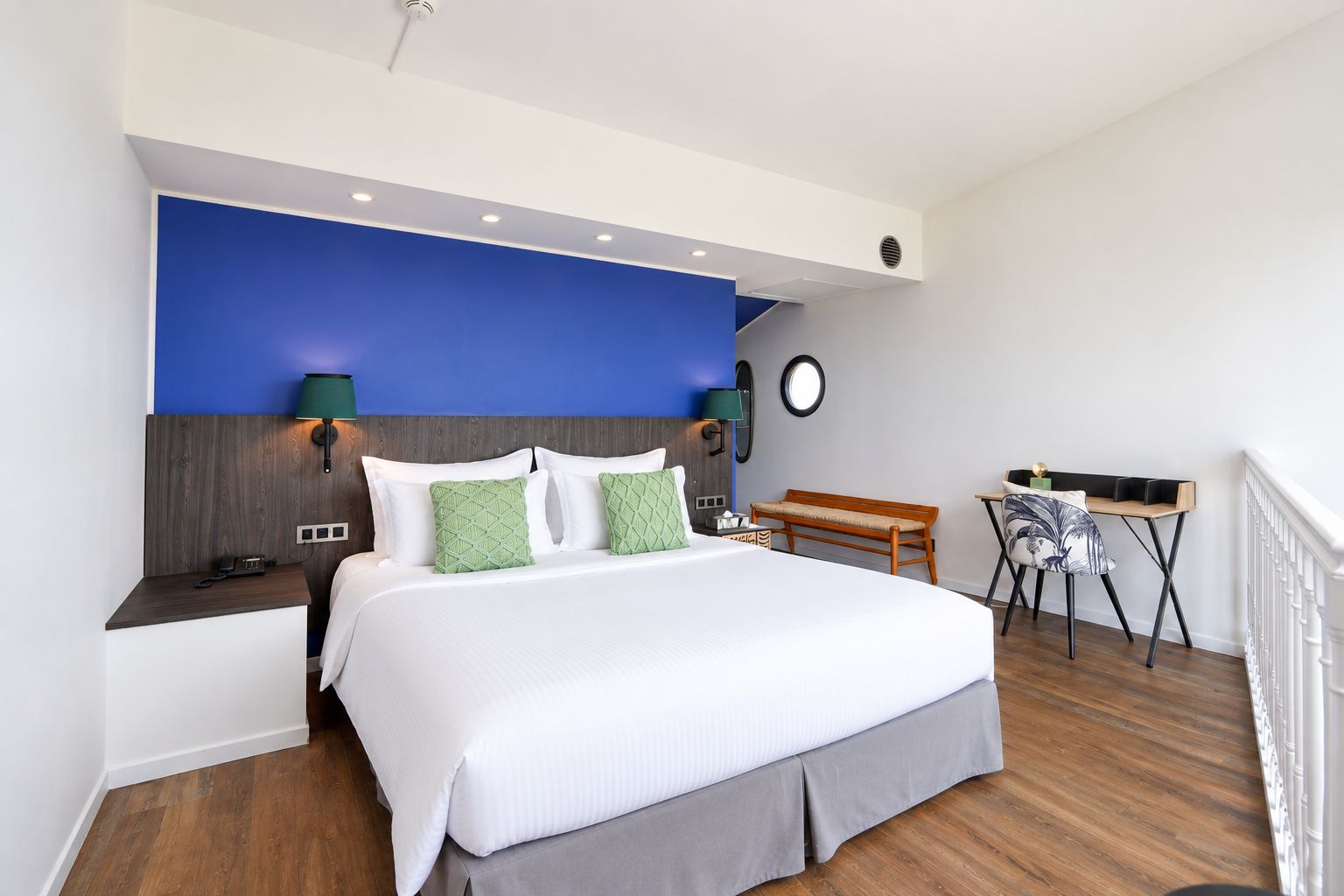 A hotel room with a large bed, a desk, a bench, and a round mirror.