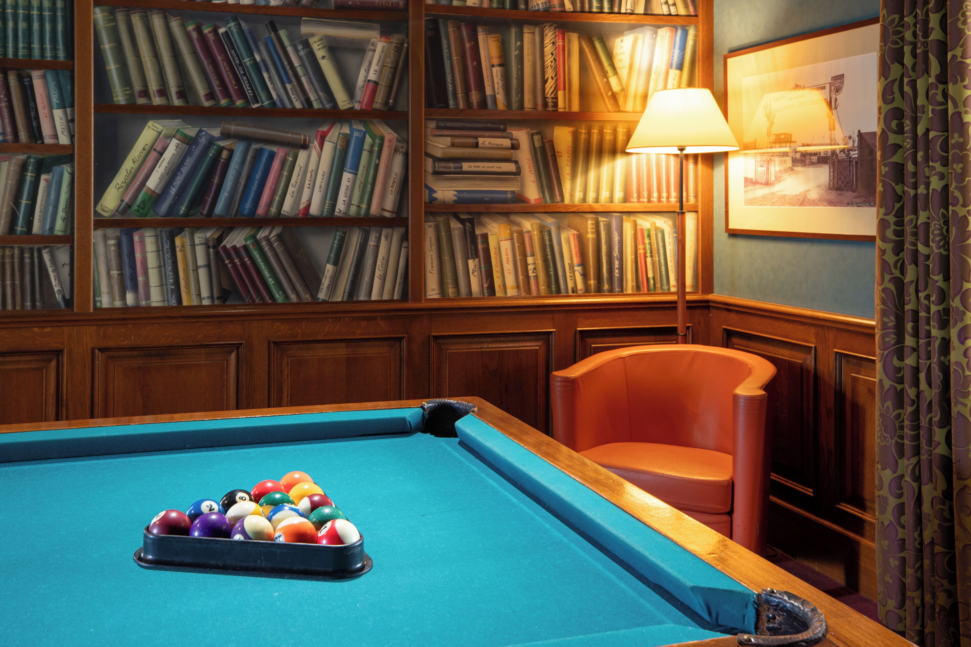 Une table de billard avec des billes, une bibliothèque remplie de livres, une lampe et un tableau encadré au mur.