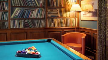 Une table de billard avec des billes, une bibliothèque remplie de livres, une lampe et un tableau encadré au mur.