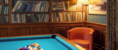 A pool table with balls, a bookshelf filled with books, a lamp, and a framed picture on the wall.