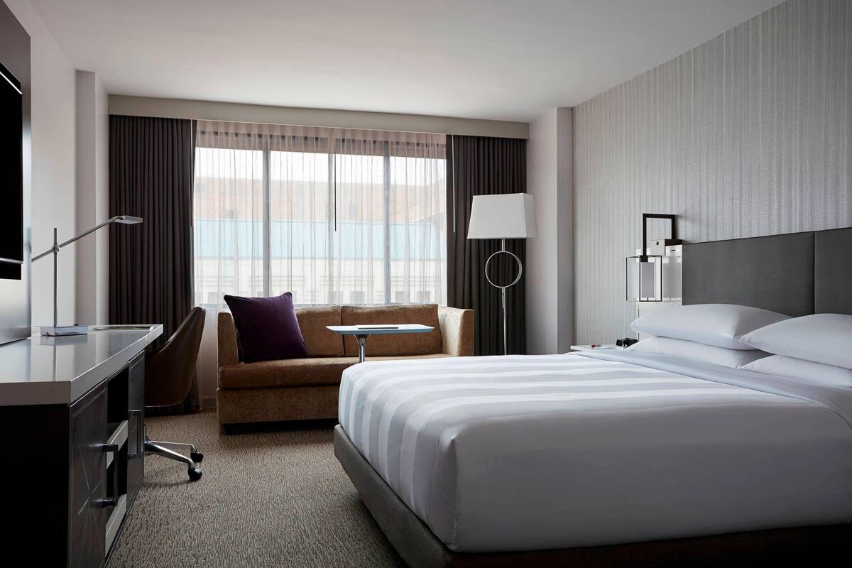 A hotel room with a bed, a desk, a chair, a small table, and a lamp.
