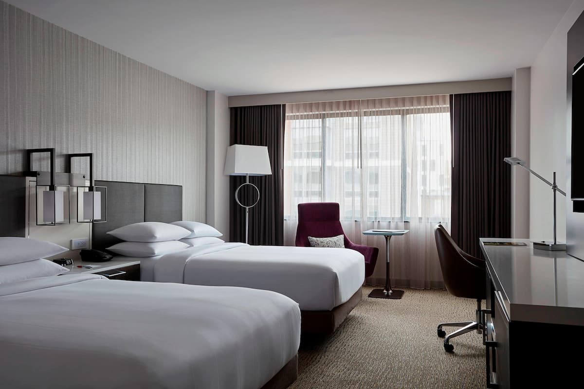 A hotel room with two beds, a desk, a chair, a lamp, and a view of the city through large windows.