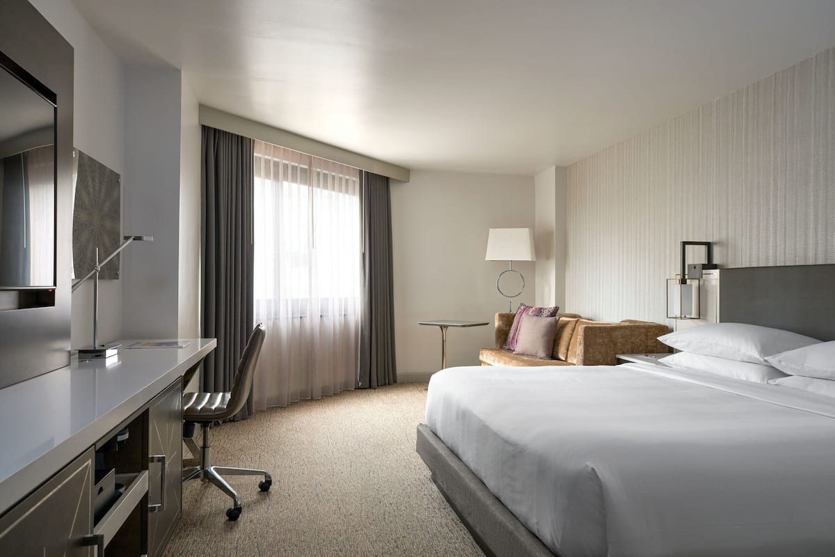 A hotel room with a bed, a desk with a chair, a small table, and a lamp.