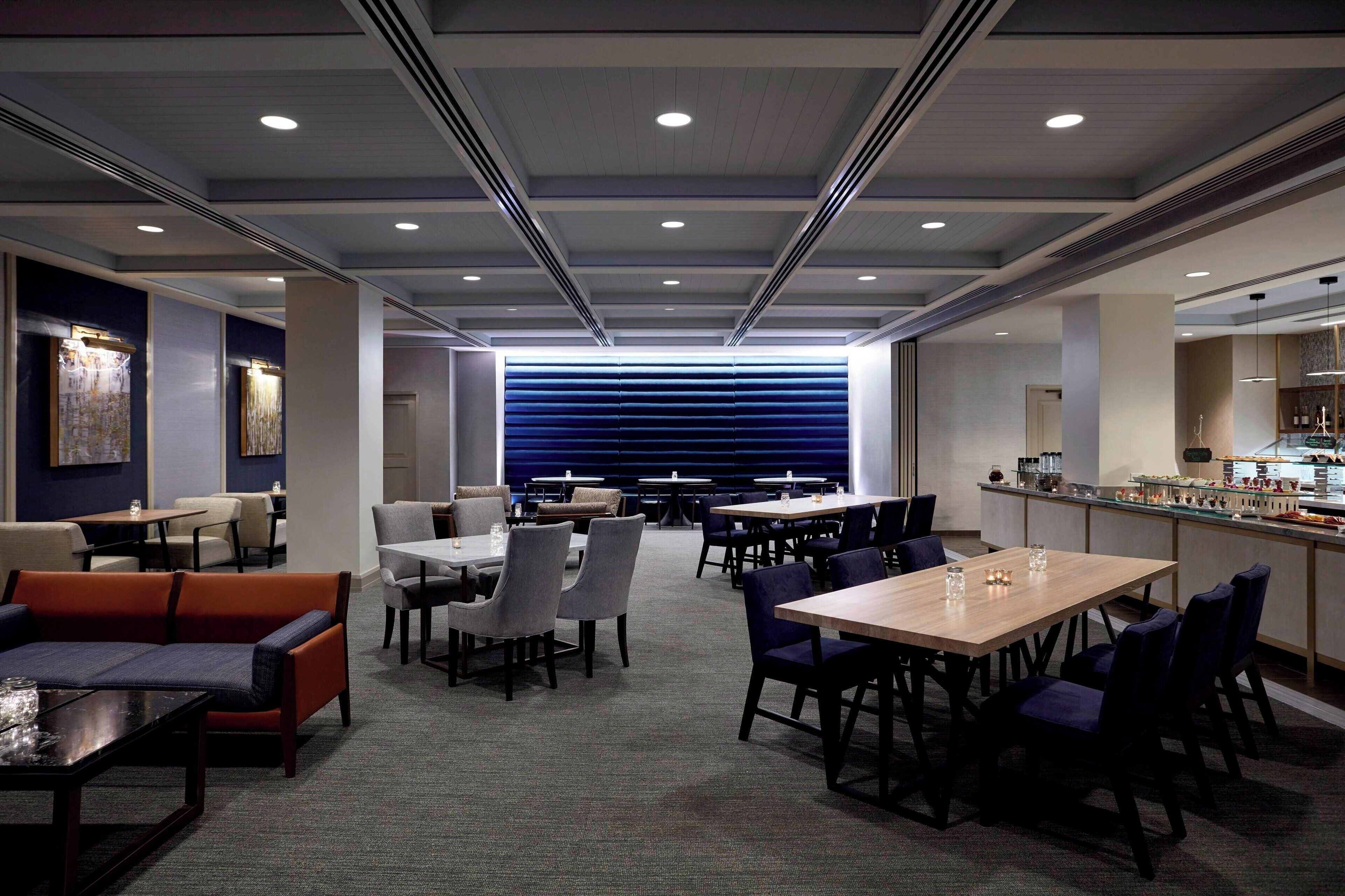 A modern hotel lobby with a bar, seating areas, and a view of the kitchen.