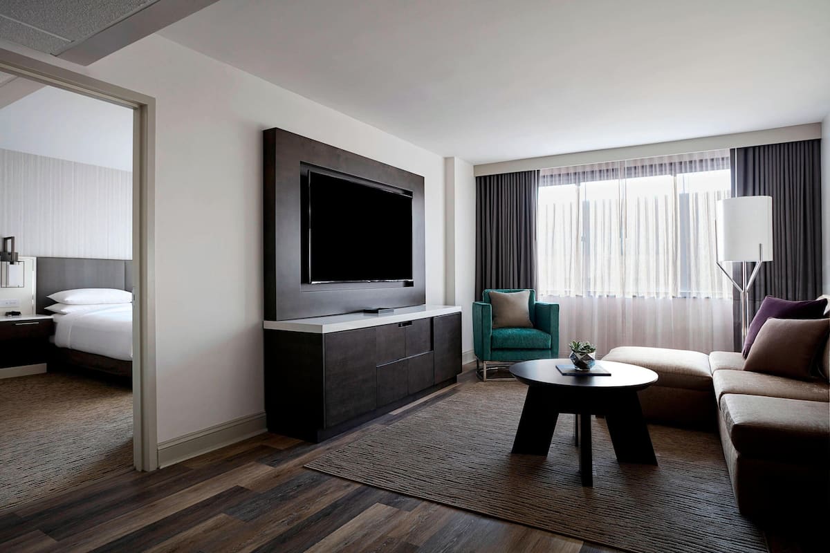 A modern hotel room with a large flat-screen TV, a sofa, a small table, and a bed with a white headboard.
