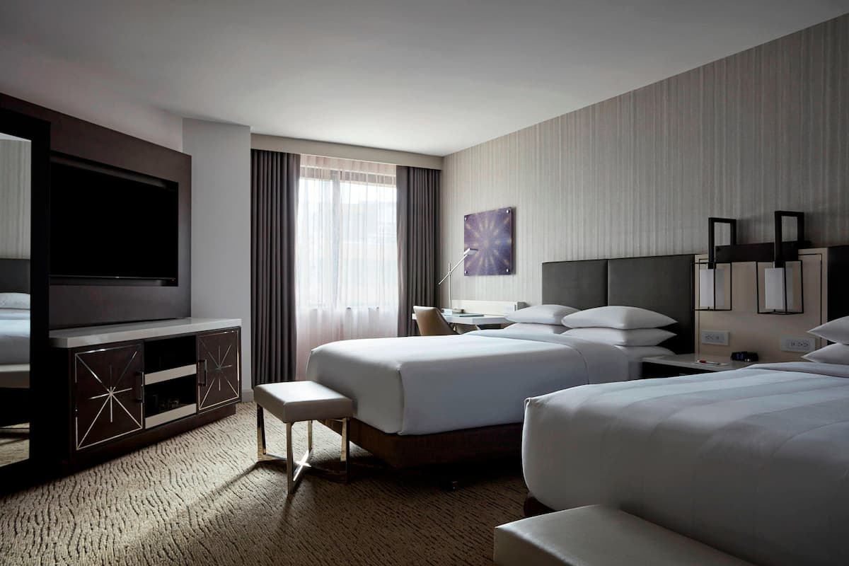 A hotel room with two beds, a TV, a desk, and a large window with curtains.