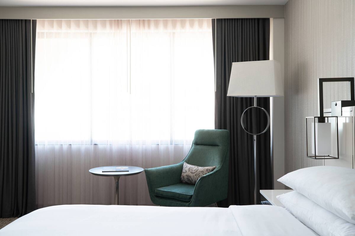 A hotel room with a bed, a green armchair, a small round table, a lamp, and a mirror.
