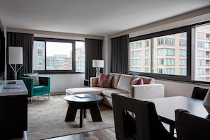 Habitación de hotel moderna con sofá, sillones, una mesa de comedor y un amplio ventanal con vistas a la ciudad.