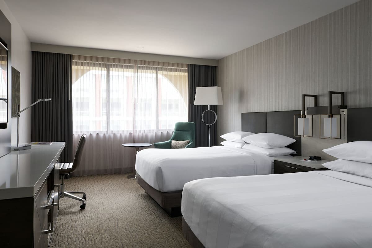 A hotel room with two beds, a desk, a chair, a lamp, and a view of the city through large windows.