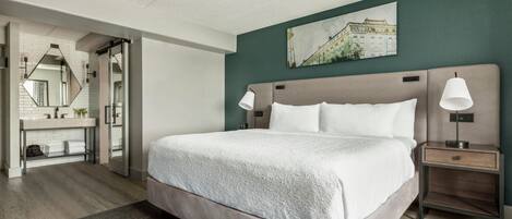 A hotel room with a bed, bedside lamps, a nightstand, a mirror, a bathroom with a sink, and a wall-mounted picture.