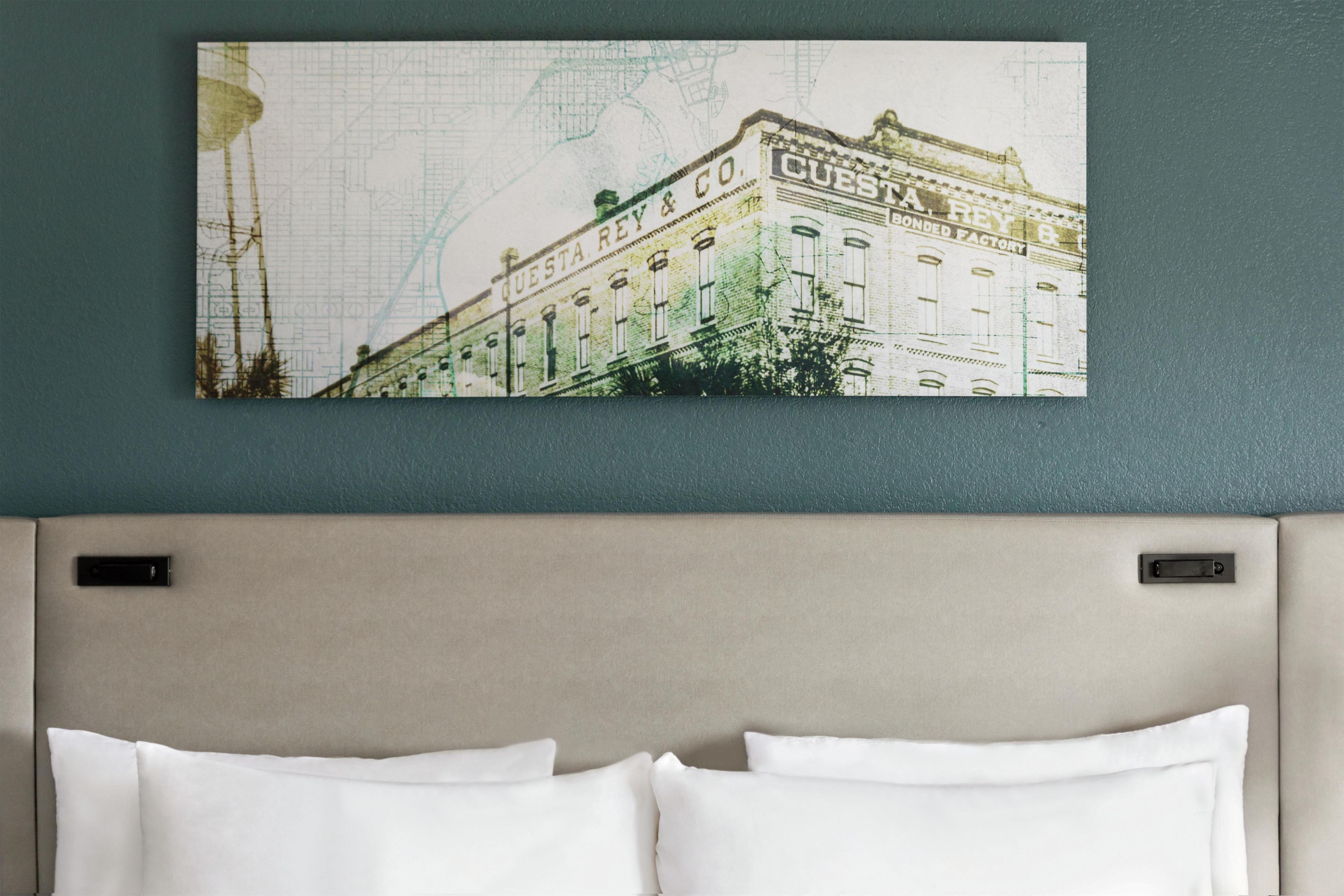 Une chambre d’hôtel avec un lit, une tête de lit et un tableau accroché au mur représentant un bâtiment, sur lequel on peut lire le texte « CUESTA REY ».