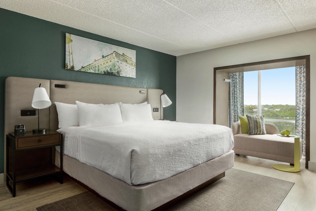 A hotel room with a large bed, bedside lamps, a nightstand, a small table, a painting on the wall, and a view through the window.