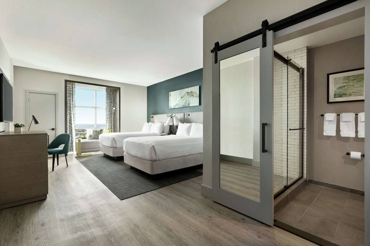 Ett modernt hotellrum med en säng, ett skrivbord, en stol och en duschhörna.