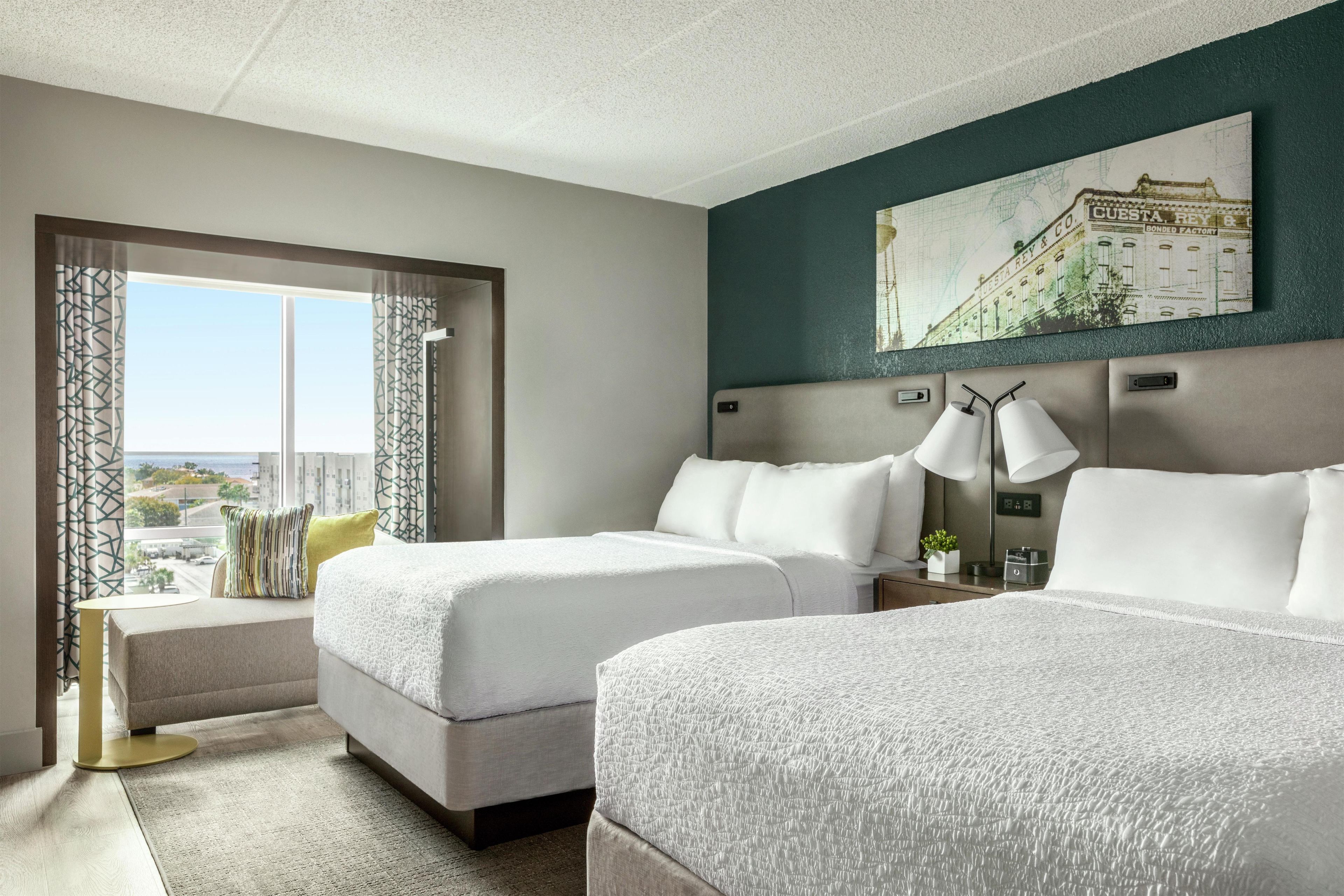 Ein Hotelzimmer mit zwei Betten, einem großen Fenster mit Vorhängen, einem Gemälde an der Wand und Blick auf die Stadt.
