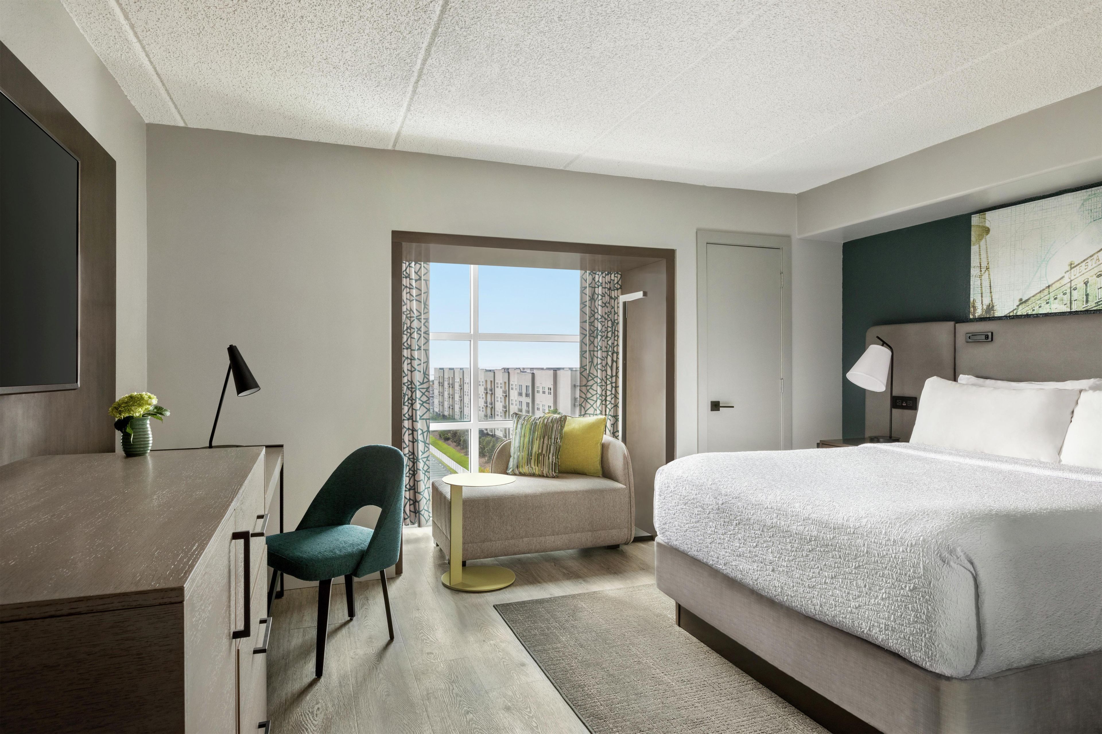 Ein Hotelzimmer mit Bett, Schreibtisch, Stuhl, Fenster mit Aussicht und einem Bild an der Wand.