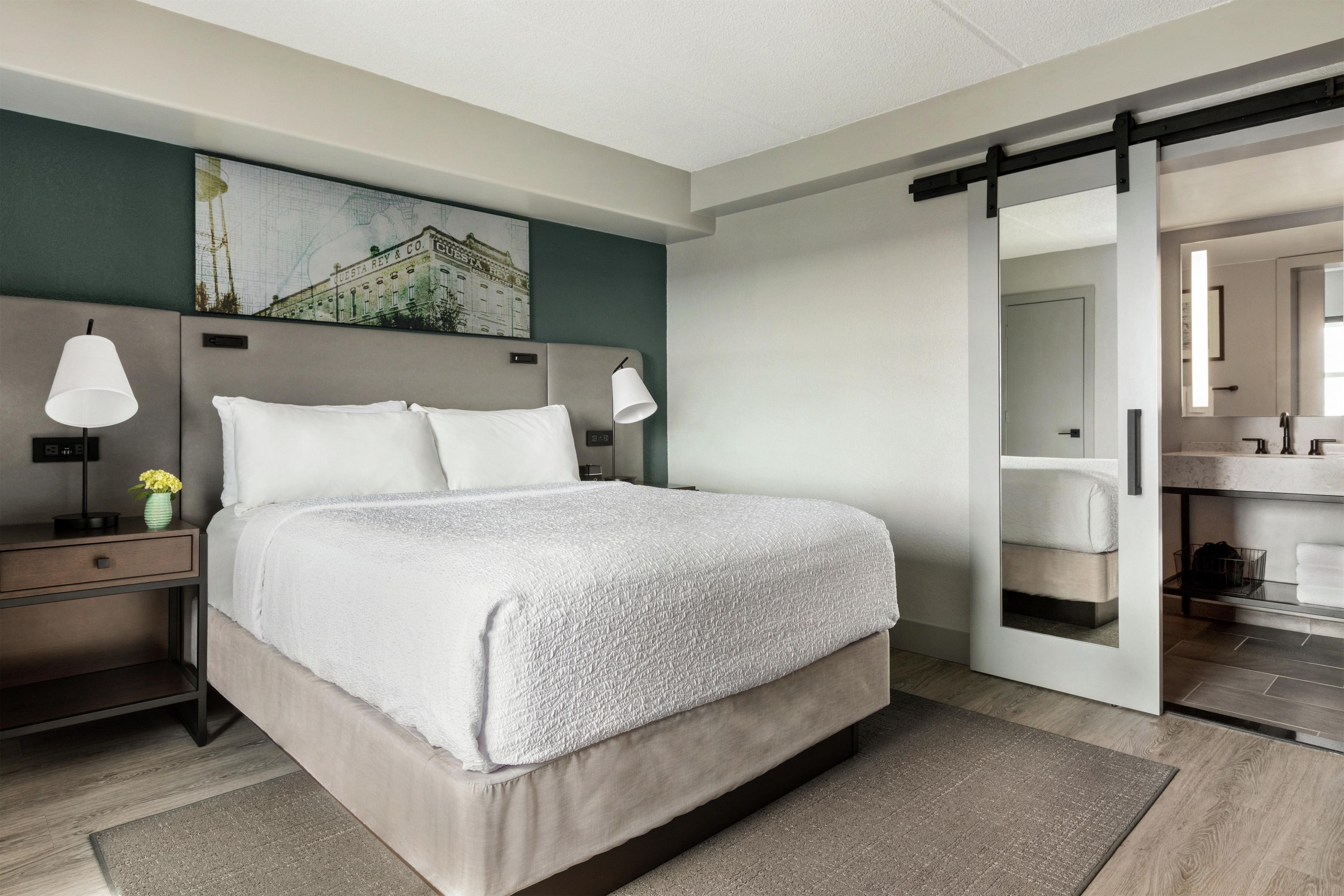 Ett modernt hotellrum med en stor säng, sänglampor, en tavla på väggen och en skjutdörr till badrummet.