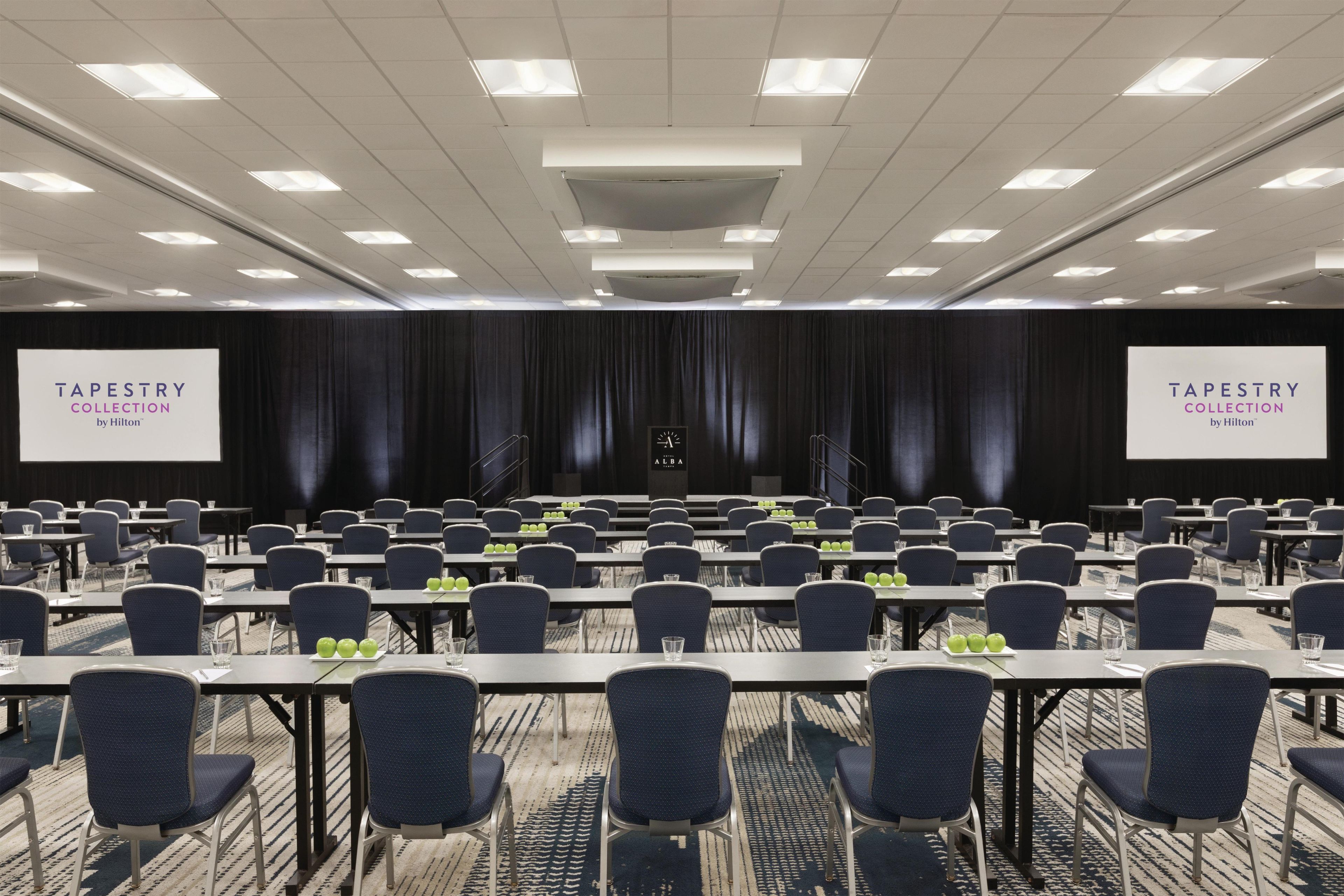 Uma sala de conferências com fileiras de mesas e cadeiras, um palco com uma tela, e dois banners com a inscrição 'TAPESTRY COLLECTION by Hilton'.