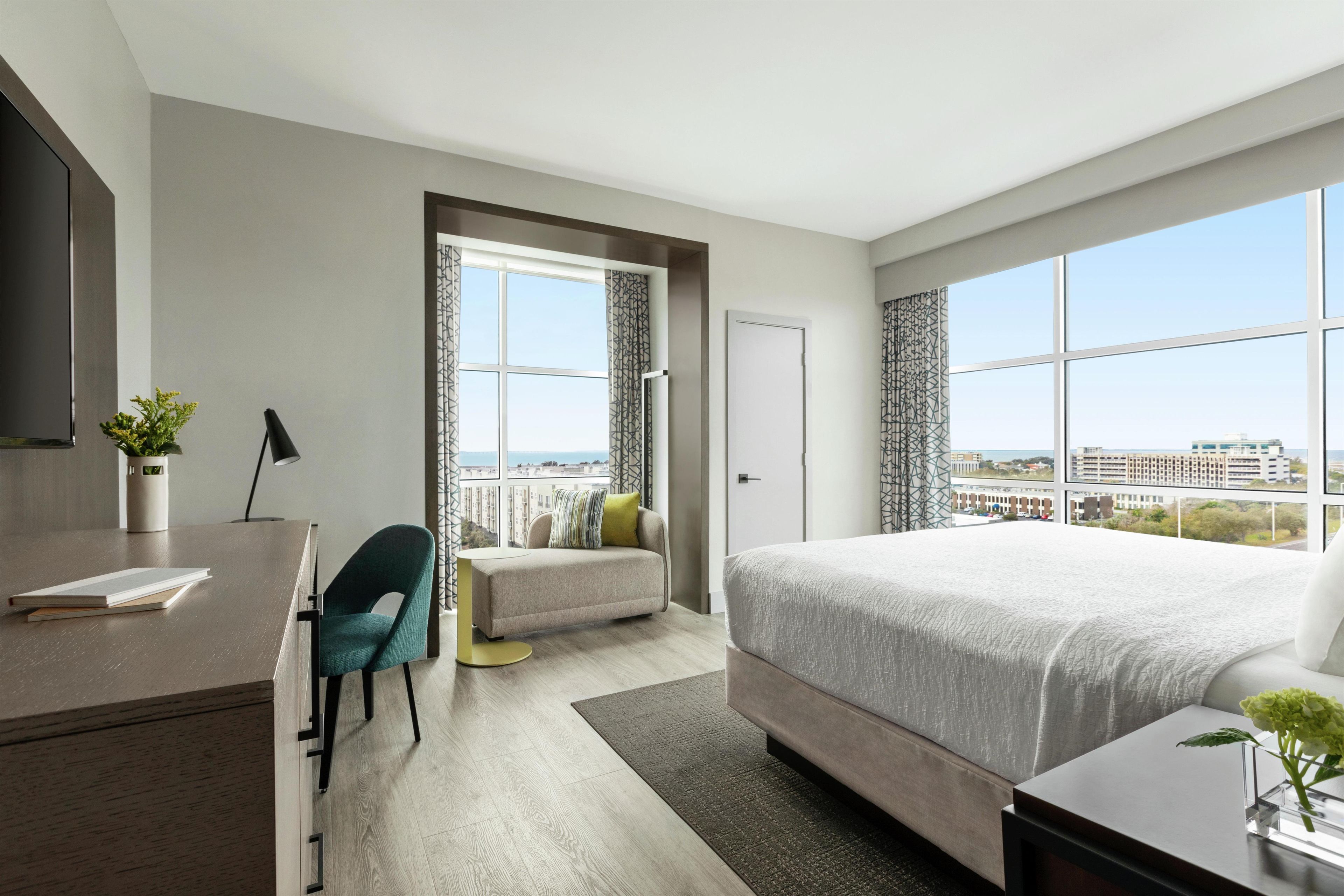Une chambre d’hôtel moderne dotée d’un grand lit, d’un bureau, d’une chaise et offrant une vue sur la ville grâce à de grandes fenêtres.