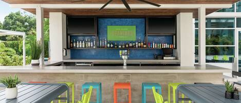 A bar area with a ceiling fan, a sign reading "Local Flavors: SPANIARD," and a variety of bottles on shelves. There are colorful stools and a table with chairs.
