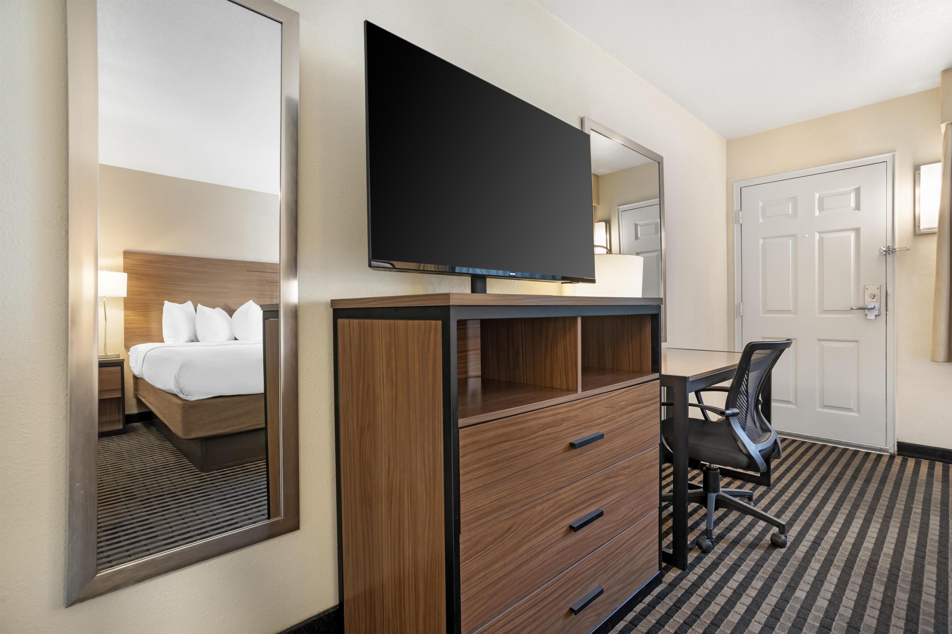 A hotel room with a bed, a desk with a chair, a flat-screen TV, a mirror, and a door.