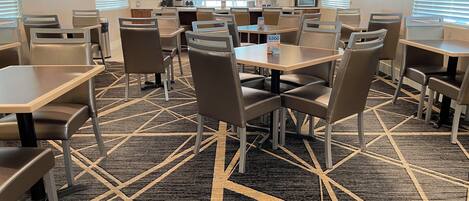 A hotel dining area with multiple tables and chairs, a patterned carpet, and artwork on the walls.