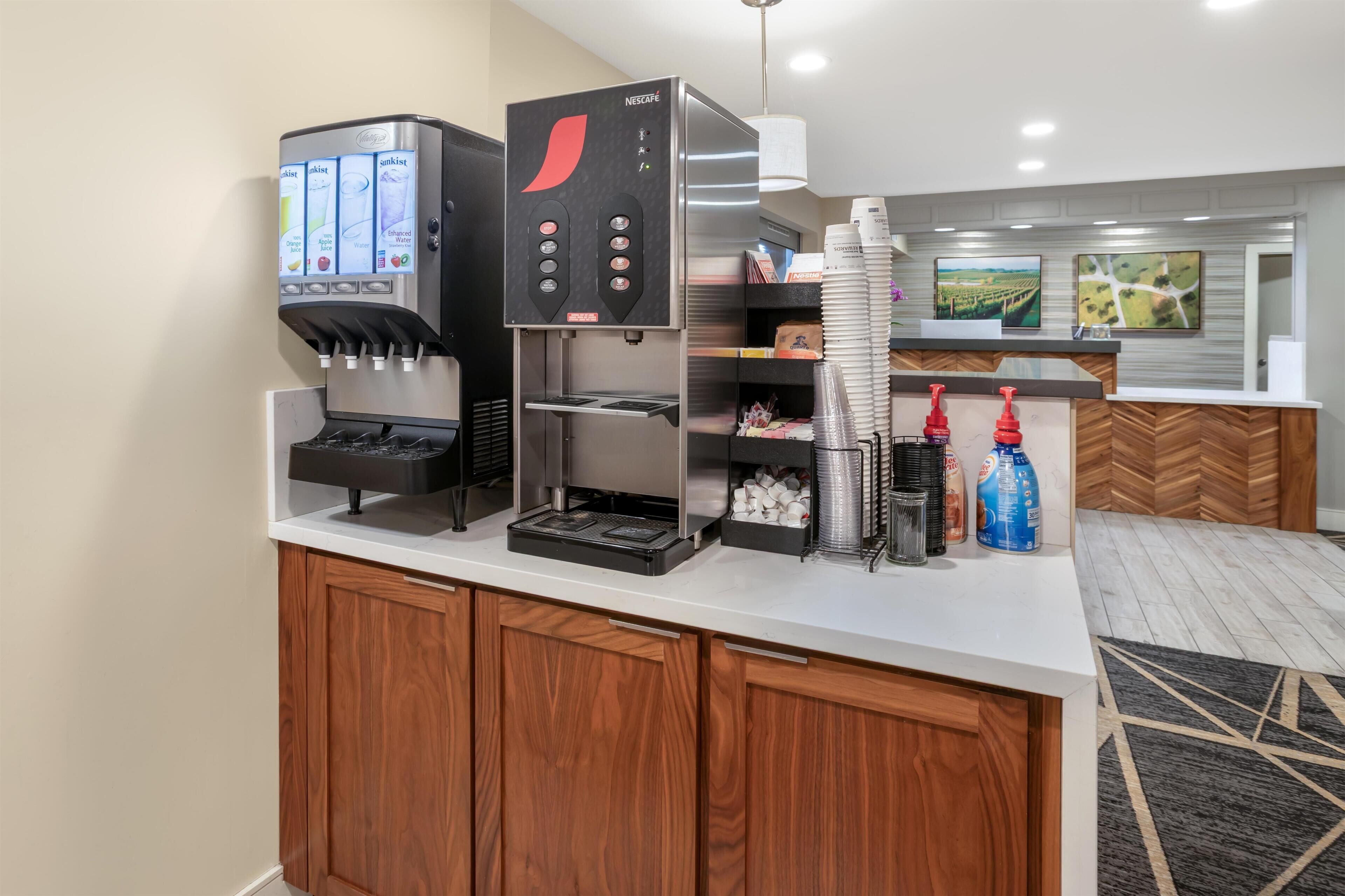 Zona de recepció d'hotel amb una màquina de cafè, un dispensador de begudes i un mostrador ple de gots i condiments.