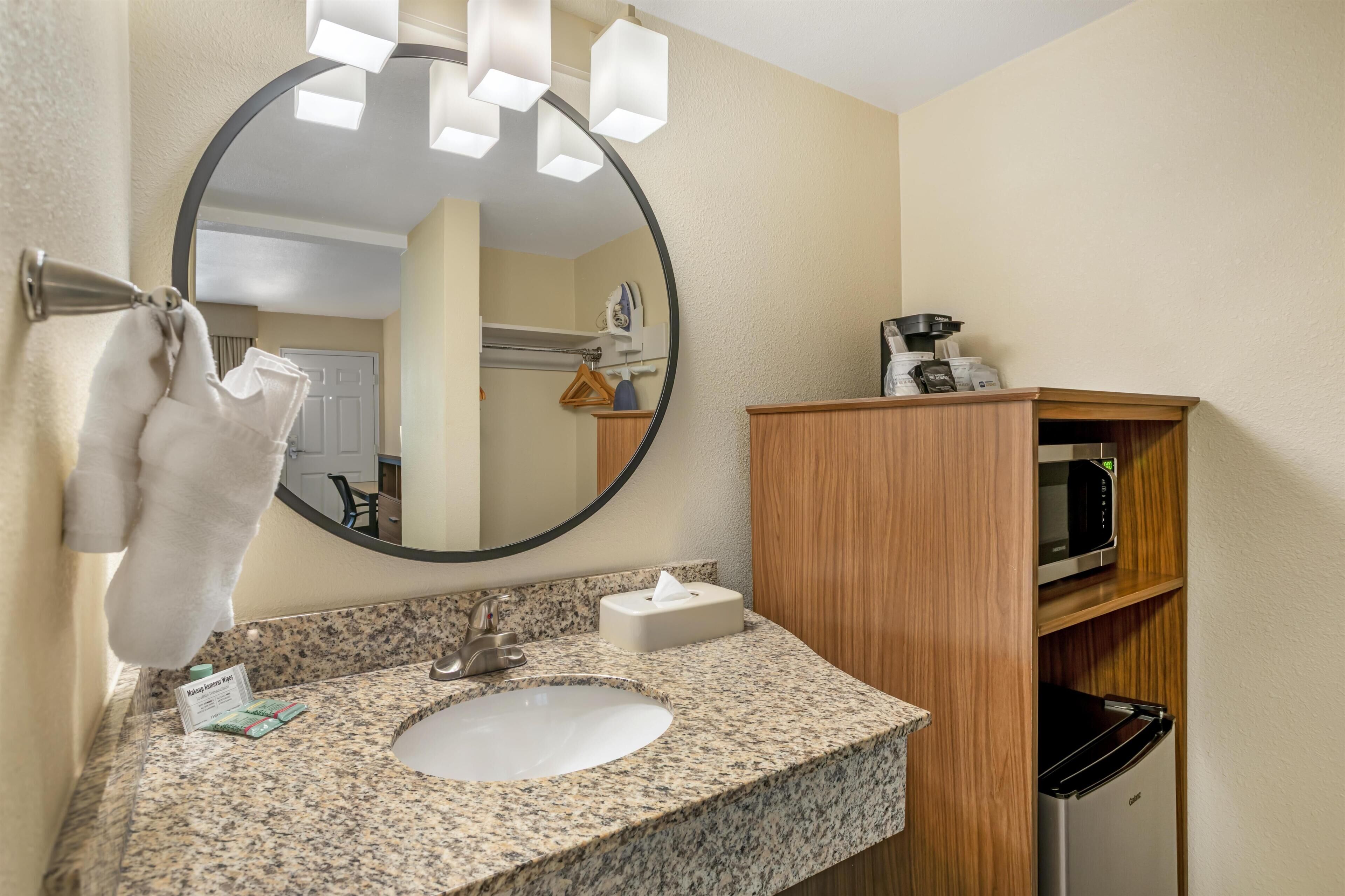 A bathroom with a granite countertop, a sink, a mirror, a towel rack, a wooden cabinet, and a microwave.