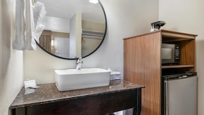 Un baño con espejo redondo, lavamanos blanco y mueble de madera con microondas y lavavajillas.