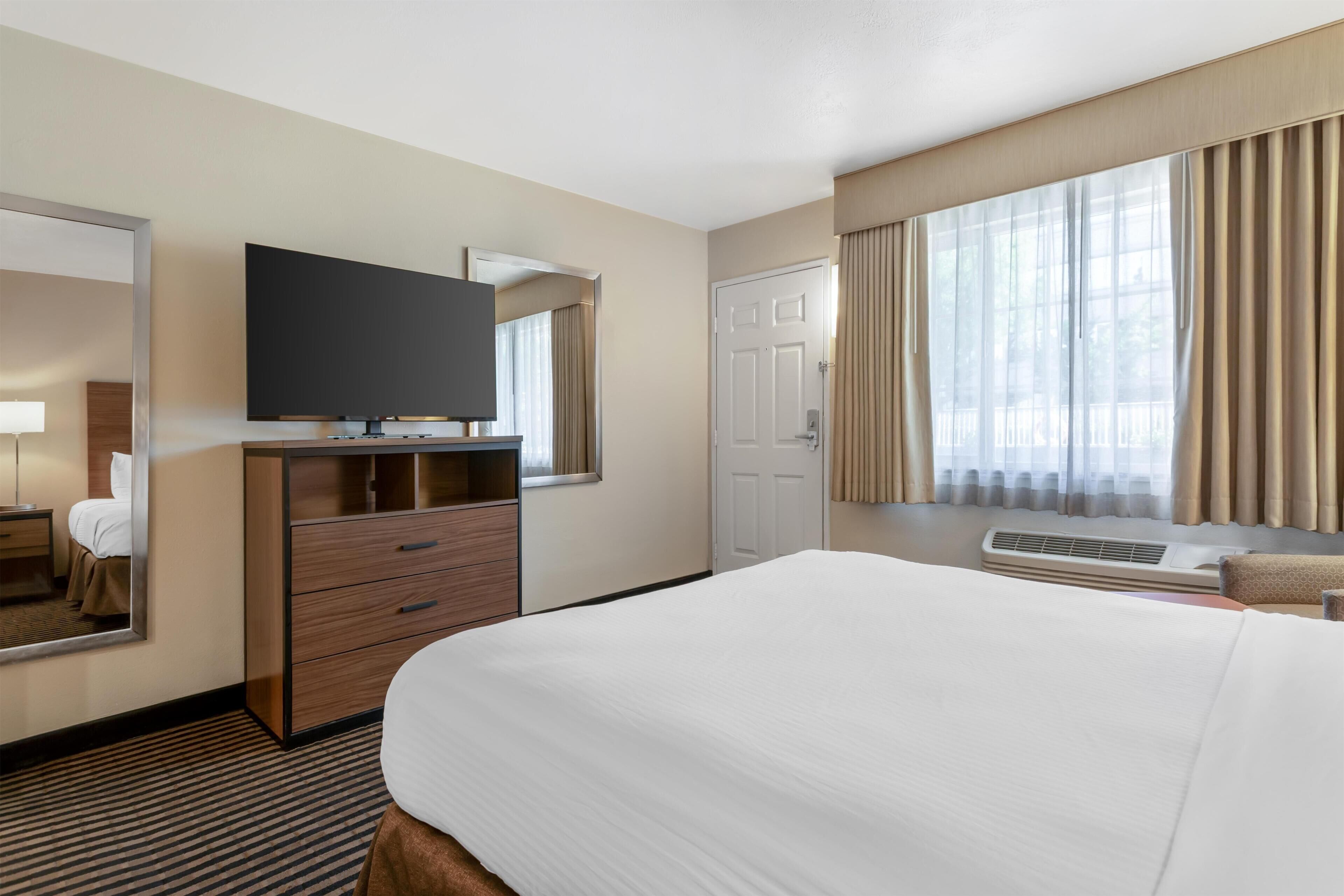 A hotel room with a large bed, a flat-screen TV, a dresser, a door, and a window with curtains.