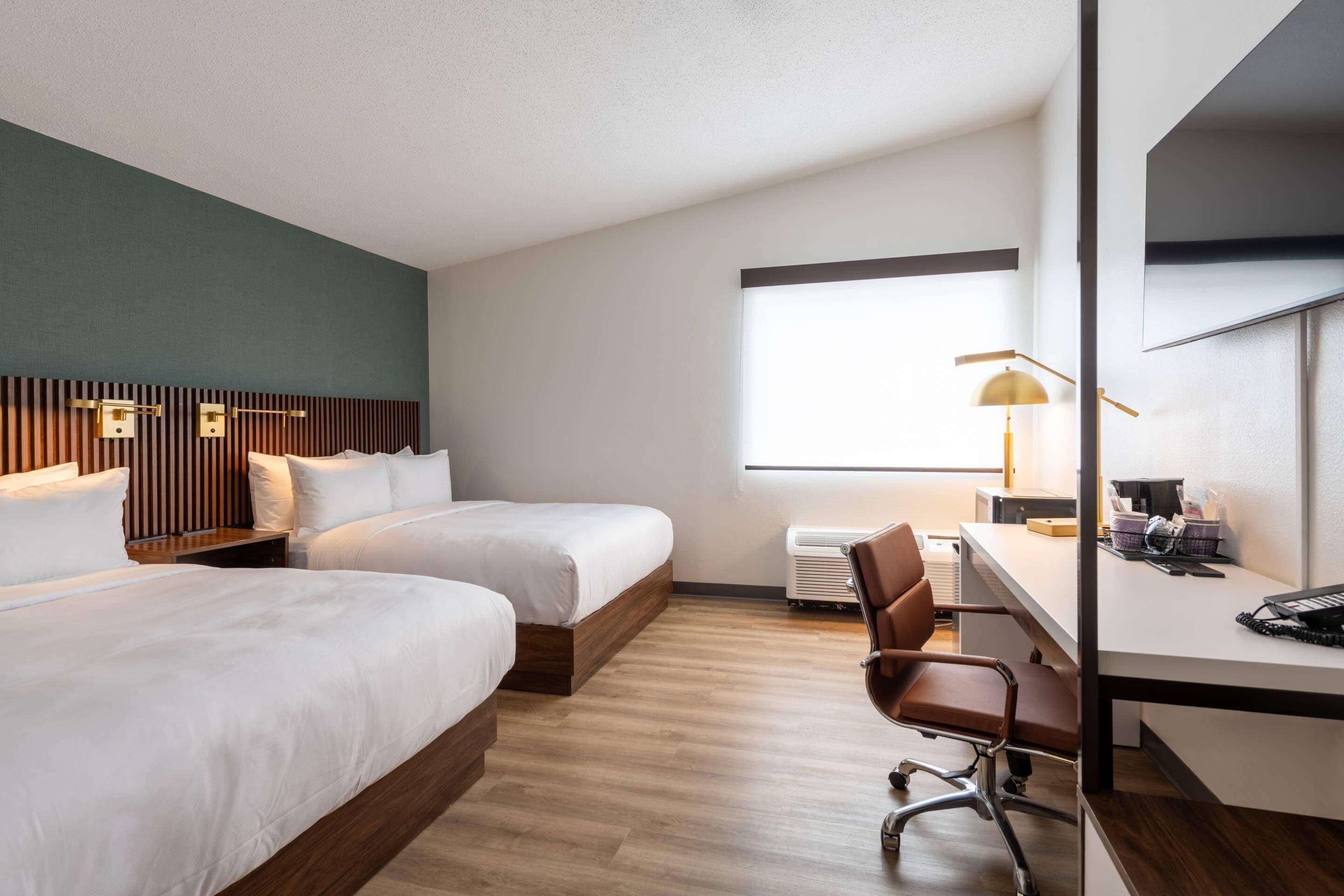 A hotel room with two beds, a desk, a chair, a lamp, and a window.